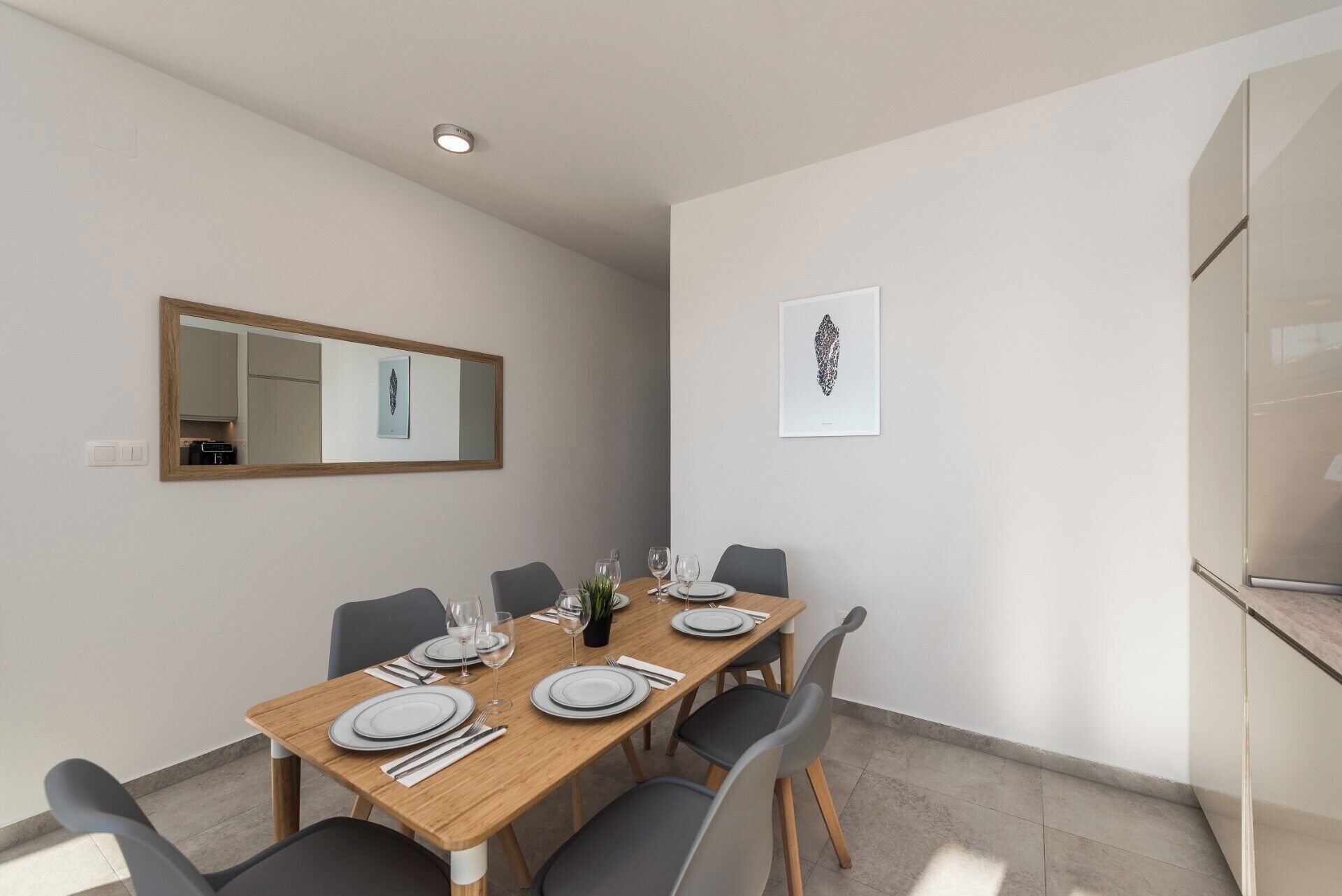 A dining area with a wooden table set for four, grey chairs, a mirror on the wall, and a framed artwork.