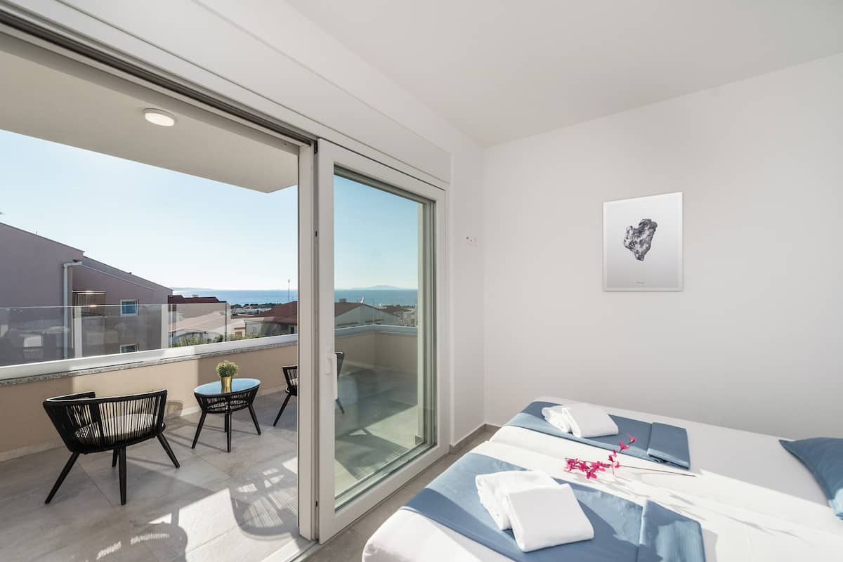 A modern hotel room with a balcony, a bed with blue bedding, a small table with a vase, and a framed picture on the wall.