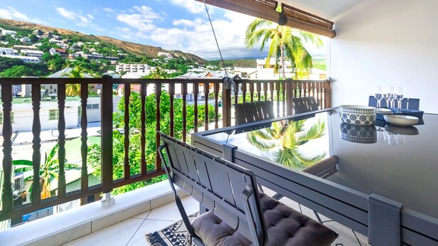 Un balcón con mesa y sillas, con vista a una zona residencial con casas y palmeras.