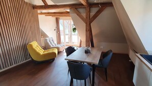 Un coin repas avec une table et des chaises en bois, un fauteuil jaune et une vue sur l’extérieur grâce à de grandes fenêtres.