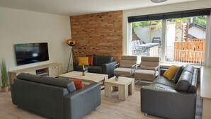 Een moderne woonkamer met een flatscreen-televisie, leren banken, houten salontafels en een groot raam met uitzicht op de omgeving.