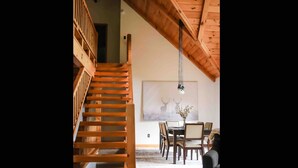 Un escalier en bois menant à l’étage supérieur, un coin repas avec une table et des chaises, et un luminaire suspendu.