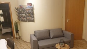 A living room with a grey sofa, a small round wooden table with a wire basket, a potted plant, and a wall-mounted magazine rack.