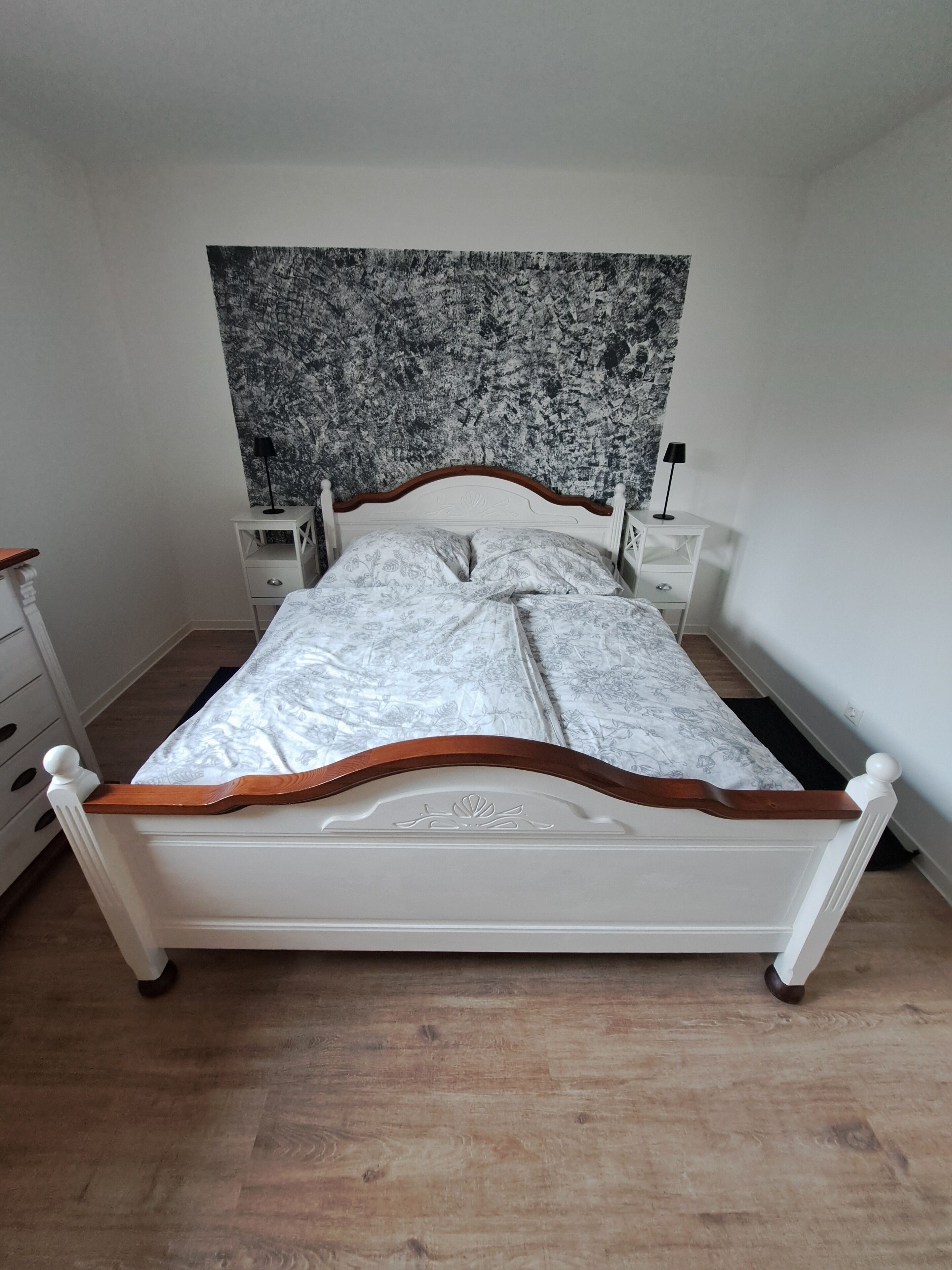 En säng med en vit ram och ett träfärgat huvudgavel, med två sängbord på var sin sida, varpå det står lampor.