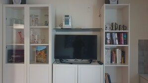 A living room with a flat-screen TV on a stand, surrounded by glass-fronted cabinets containing books, decorative items, and kitchenware.