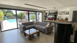 A modern living room with a sectional sofa, a coffee table, and a kitchen area visible through glass doors. - ANGLET, 8 pers COAST HOUSE WITH SWIMMING POOL, NEAR THE BEACH, cycle path, GOLF (Anglet)