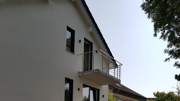 Un bâtiment moderne à deux étages, doté d’un balcon, d’une allée pavée et entouré de verdure.