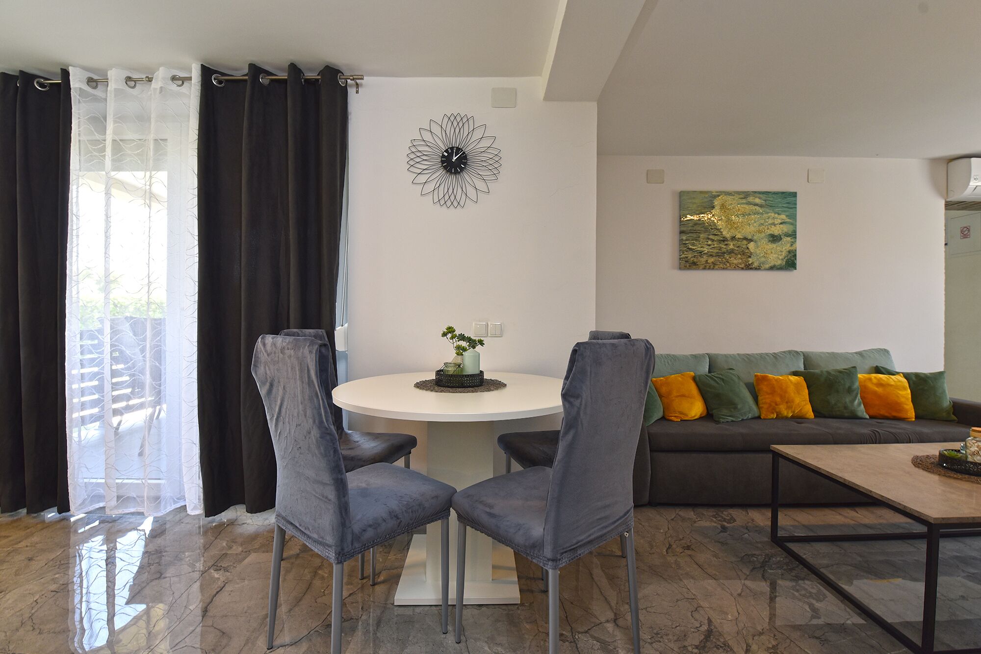 Ein modernes Wohnzimmer mit einem runden Esstisch, zwei grauen Polsterstühlen, einem grauen Sofa mit bunten Kissen, einer dekorativen Wanduhr und einem Gemälde.