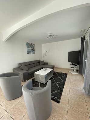 Uma sala de estar moderna com um sofá modular cinzento, uma mesa de centro de vidro e duas poltronas cinzentas. Uma televisão de tela plana está fixada na parede, e há uma planta decorativa sobre a mesa de centro.