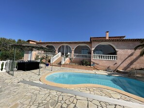 Uma área de piscina com um terraço de pedra, um espaço de estar coberto e um edifício com janelas em arco e varanda.