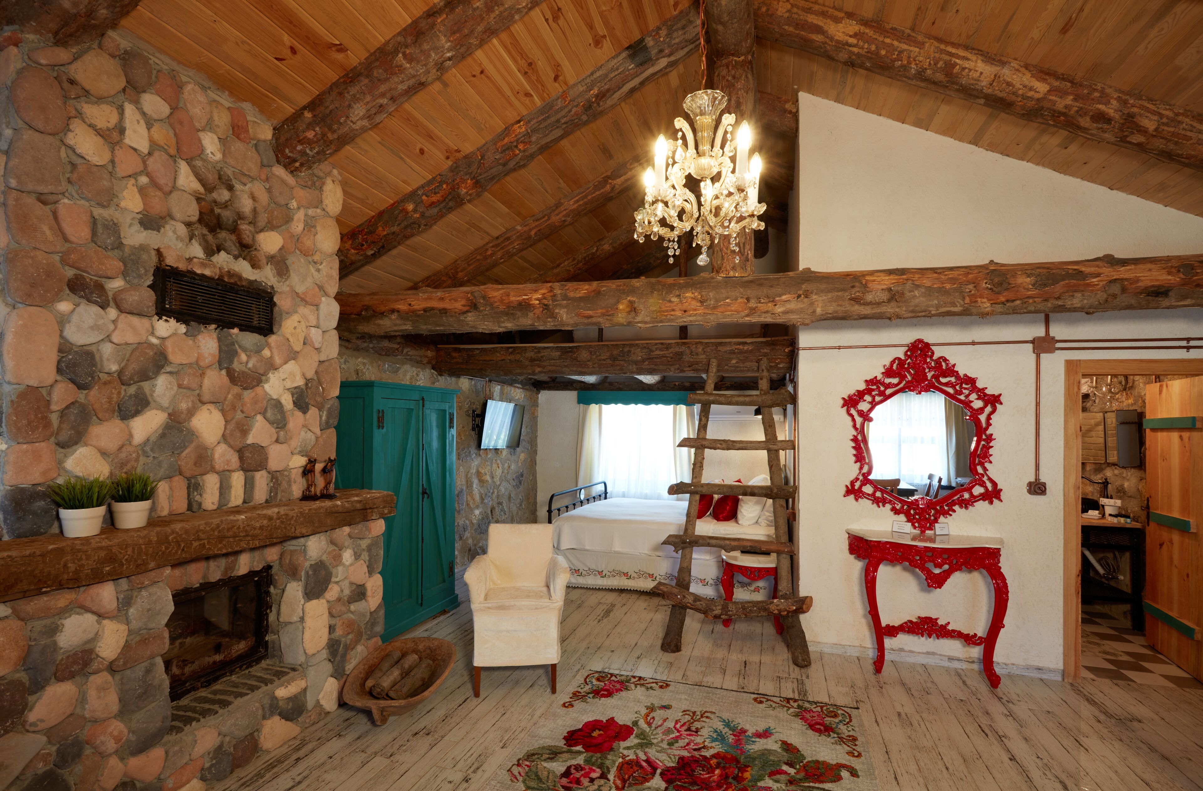 A rustic room with a stone fireplace, wooden beams, a chandelier, a wooden ladder, a red mirror, and a bed with white bedding.