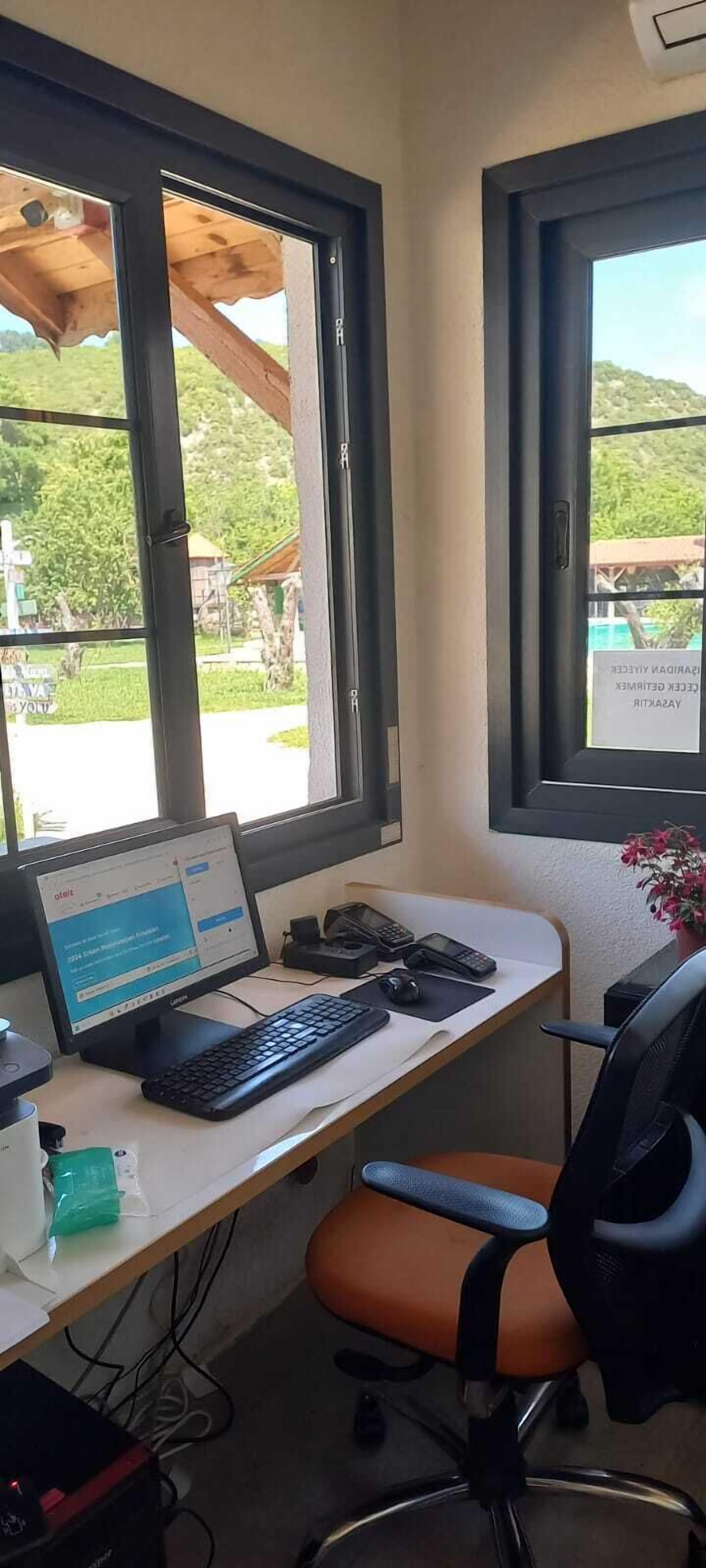 Un espacio de trabajo con una computadora portátil, teclado y ratón sobre un escritorio, una silla y una ventana que ofrece vistas a zonas verdes y un edificio.