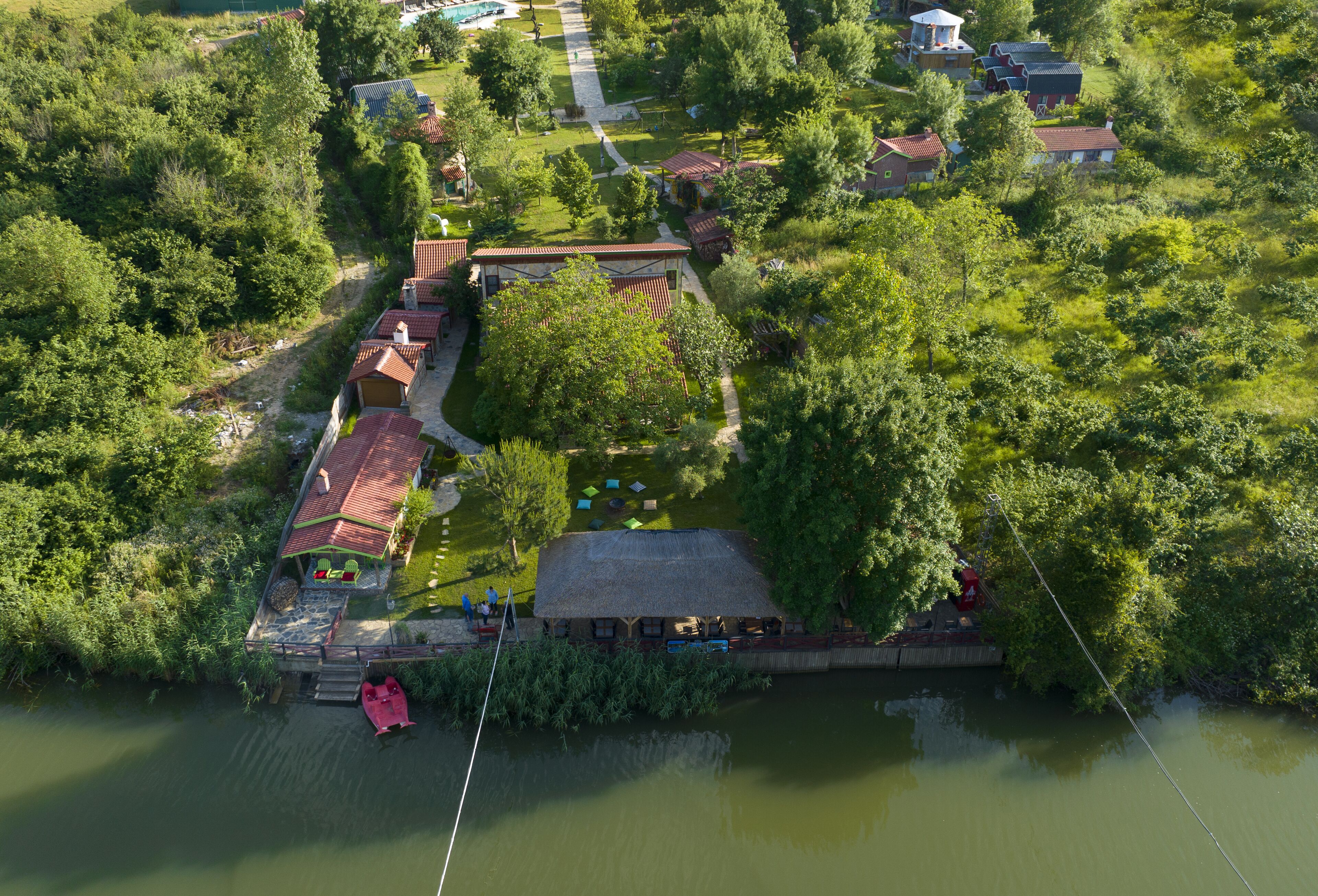 An aerial view of a riverside property with a thatched-roof building, a dock with a red boat, and a small bridge connecting to the main area.