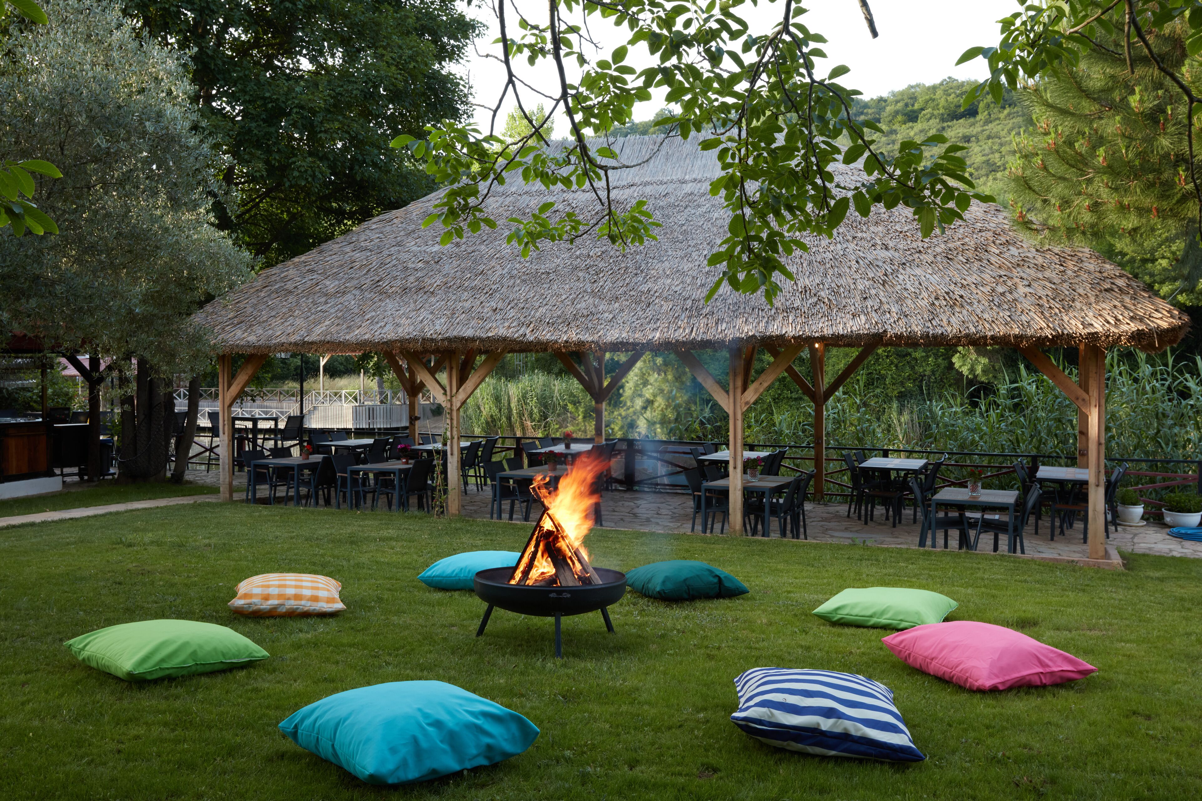 Un área de comedor al aire libre con techo de paja y una chimenea rodeada de cojines de colores.