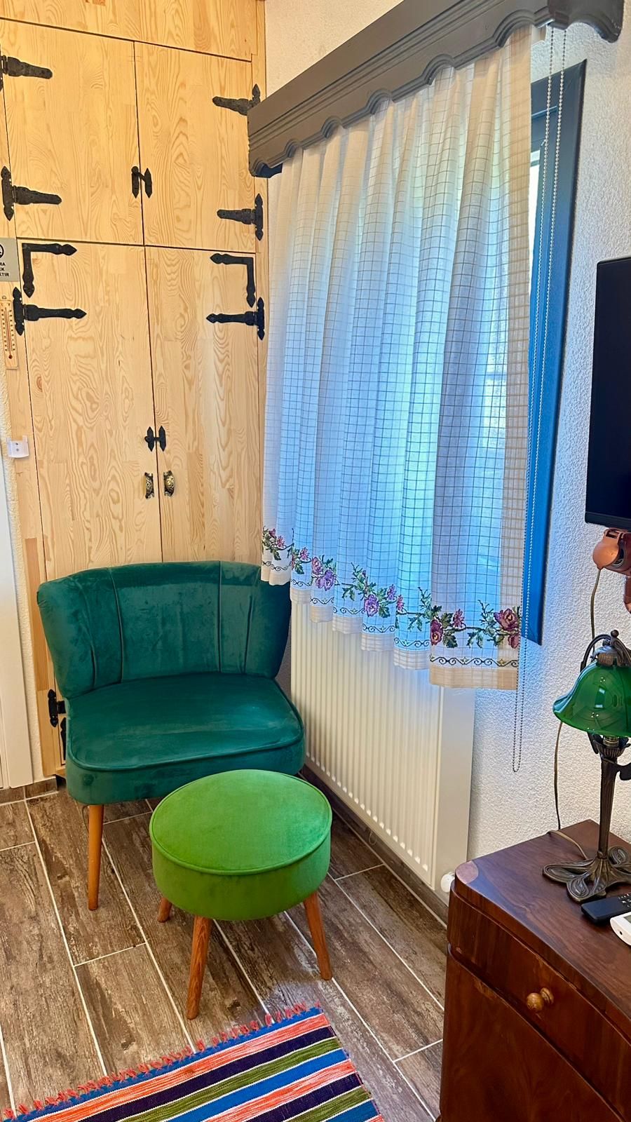 Huoneessa on vihreä sohva, samanvärinen jalkarahi, puinen kaappi, patteri ja ikkuna, jossa on verho.