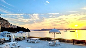 A beachside patio with picnic tables and umbrellas, overlooking a calm sea and distant mountains during sunset. - Yeosu Arens Pension (Yeosu)