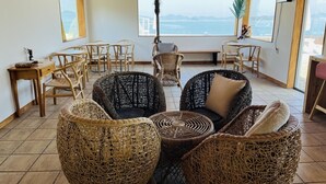 A spacious room with wicker furniture, a large window offering a view of the sea, and a ceiling with wooden beams. - Yeosu Arens Pension (Yeosu)