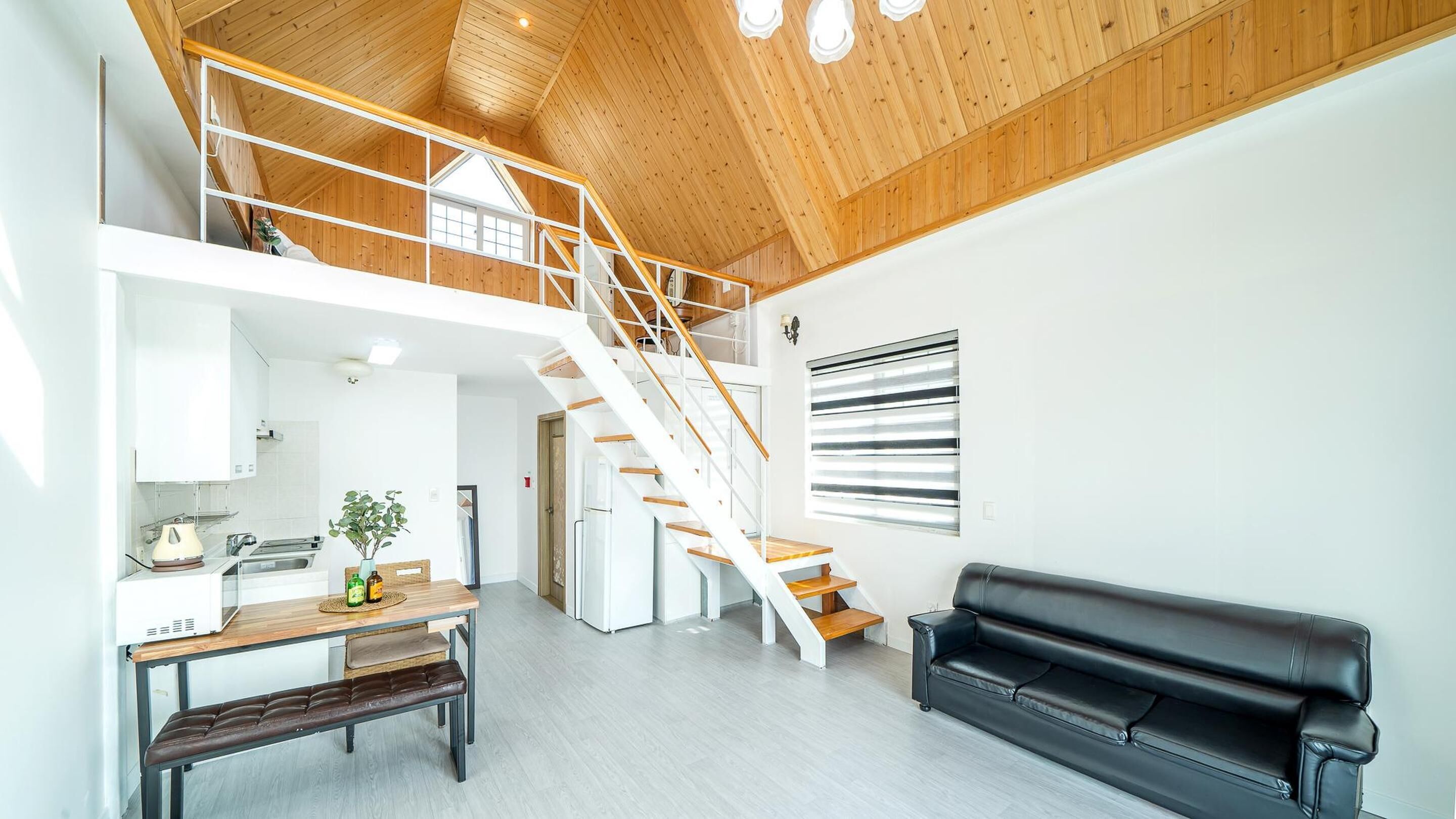 A spacious living area with a wooden staircase, a black leather sofa, and a kitchen area with a dining table and bench.