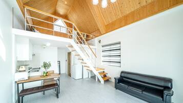 A spacious living area with a wooden staircase, a black leather sofa, and a kitchen area with a dining table and bench.