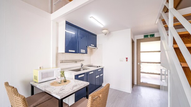 A modern kitchen with dark blue cabinets, a white countertop, and a microwave. There is a dining area with a table and chairs, and a staircase leading to an upper level. - Yeosu Arens Pension (Yeosu)