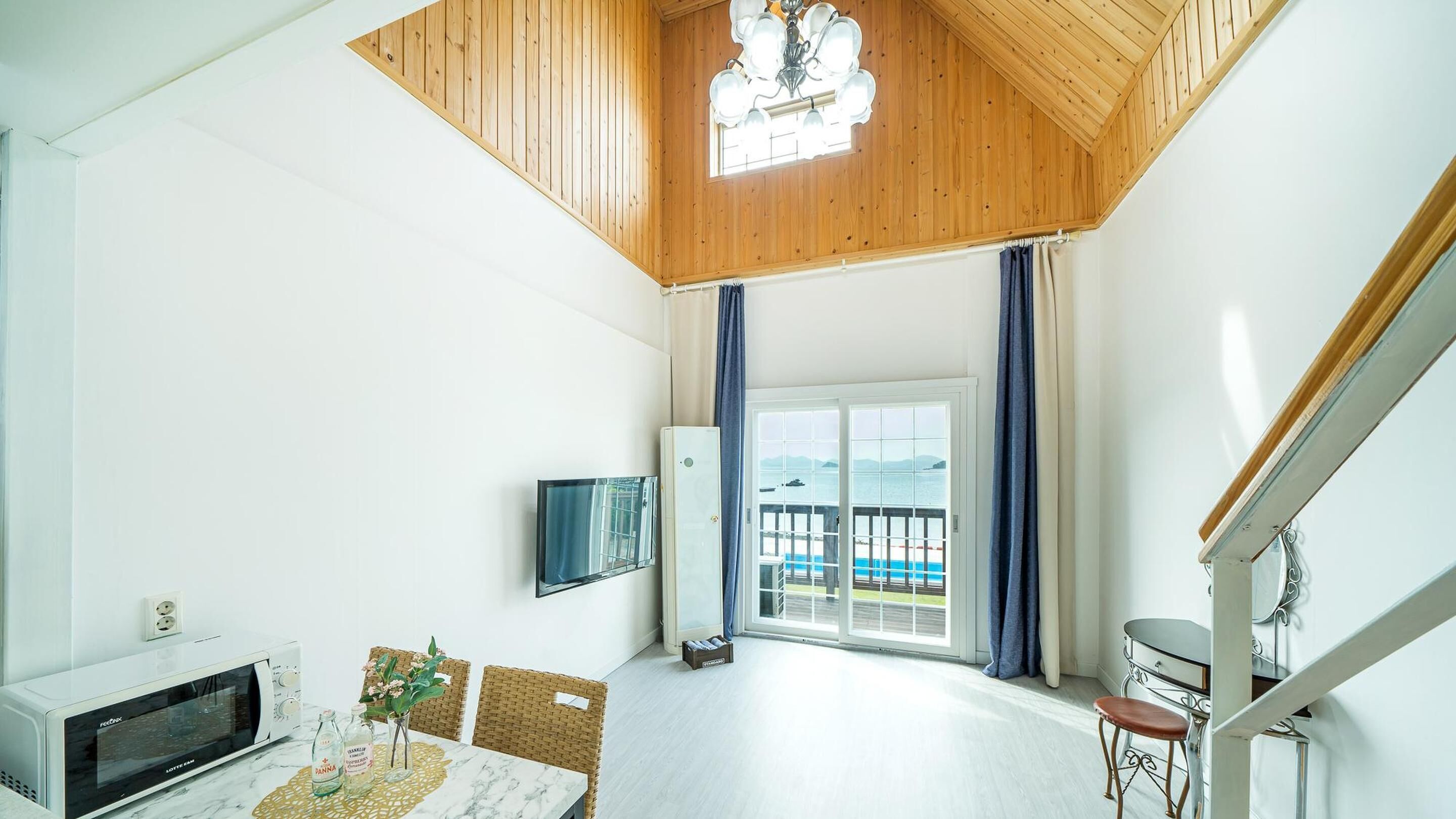 A room with a wooden ceiling, a television mounted on the wall, a dining table with chairs, and a view of the sea through a glass door.