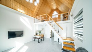 A spacious interior with a high wooden ceiling, a staircase leading to an upper level, a dining area with a table and chairs, and a kitchen visible in the background. - Yeosu Arens Pension (Yeosu)