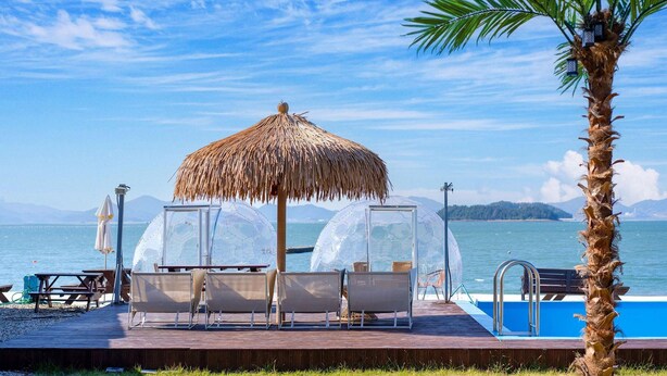 A beachside pool area with a thatched umbrella, transparent dome structures, and wooden picnic tables. - Yeosu Arens Pension (Yeosu)