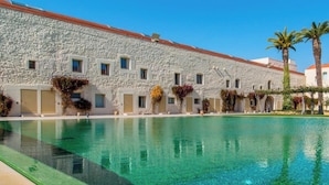 A swimming pool with clear water, a stone building with multiple windows, and a palm tree. - Convento das Bernardas Lux T2 (Tavira)