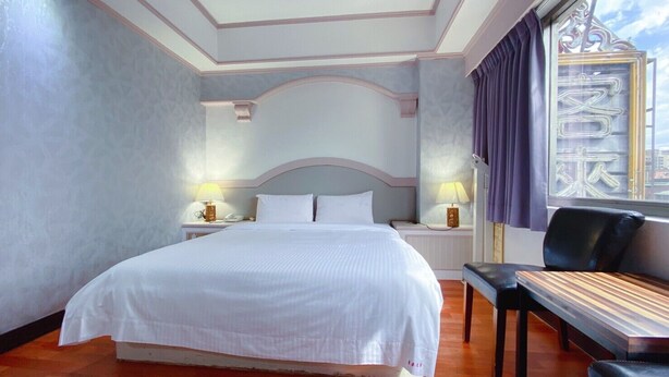 A hotel room with a large bed, two bedside tables with lamps, a window with curtains, and a bench.