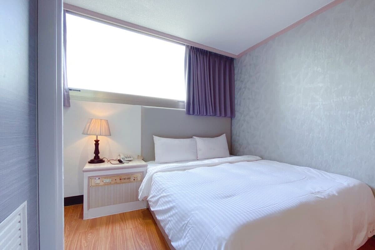 A hotel room with a bed, a bedside table with a lamp, a phone, and a window with a curtain.