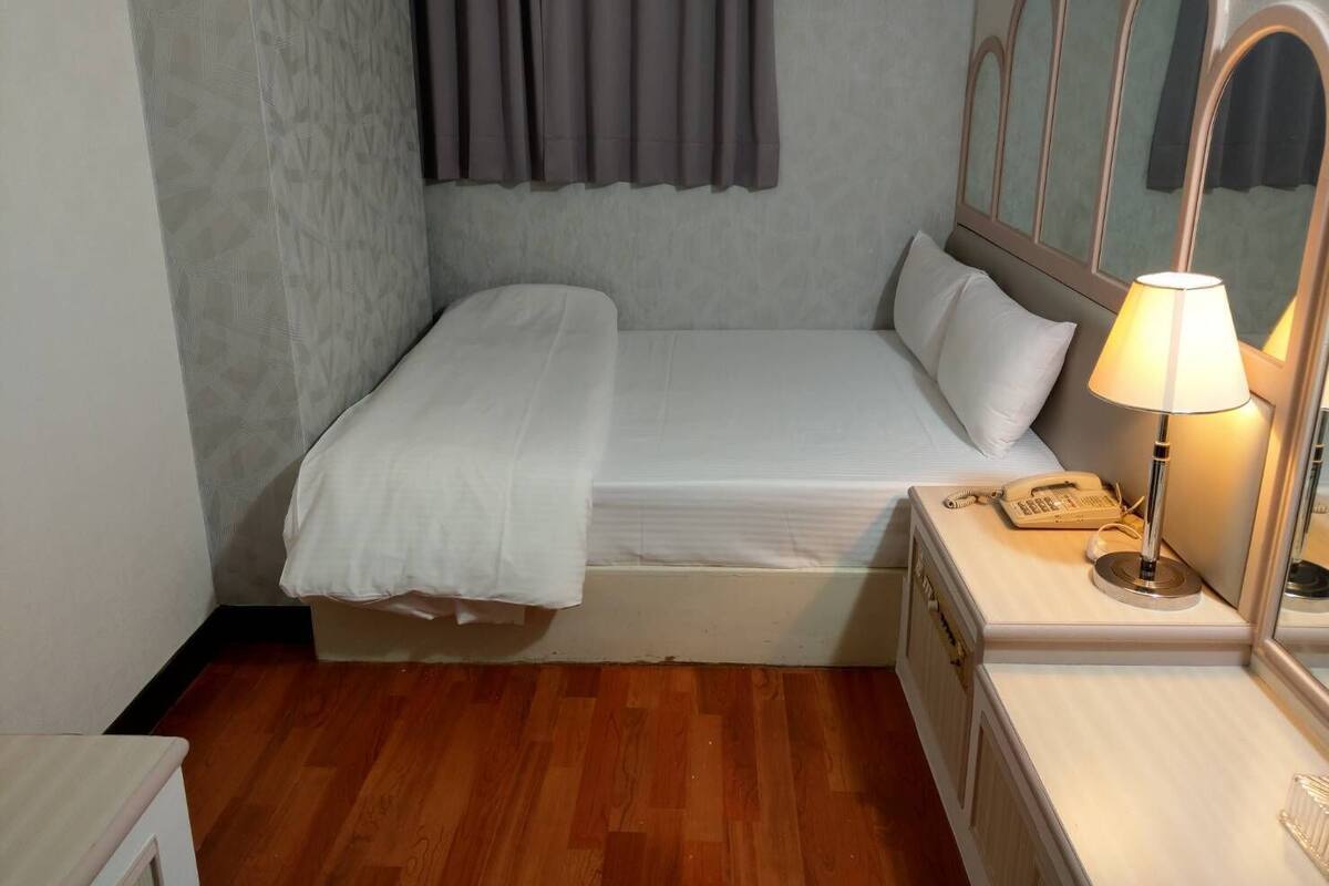 A single bed with a white mattress and pillows, a bedside table with a lamp, and a small bedside telephone.
