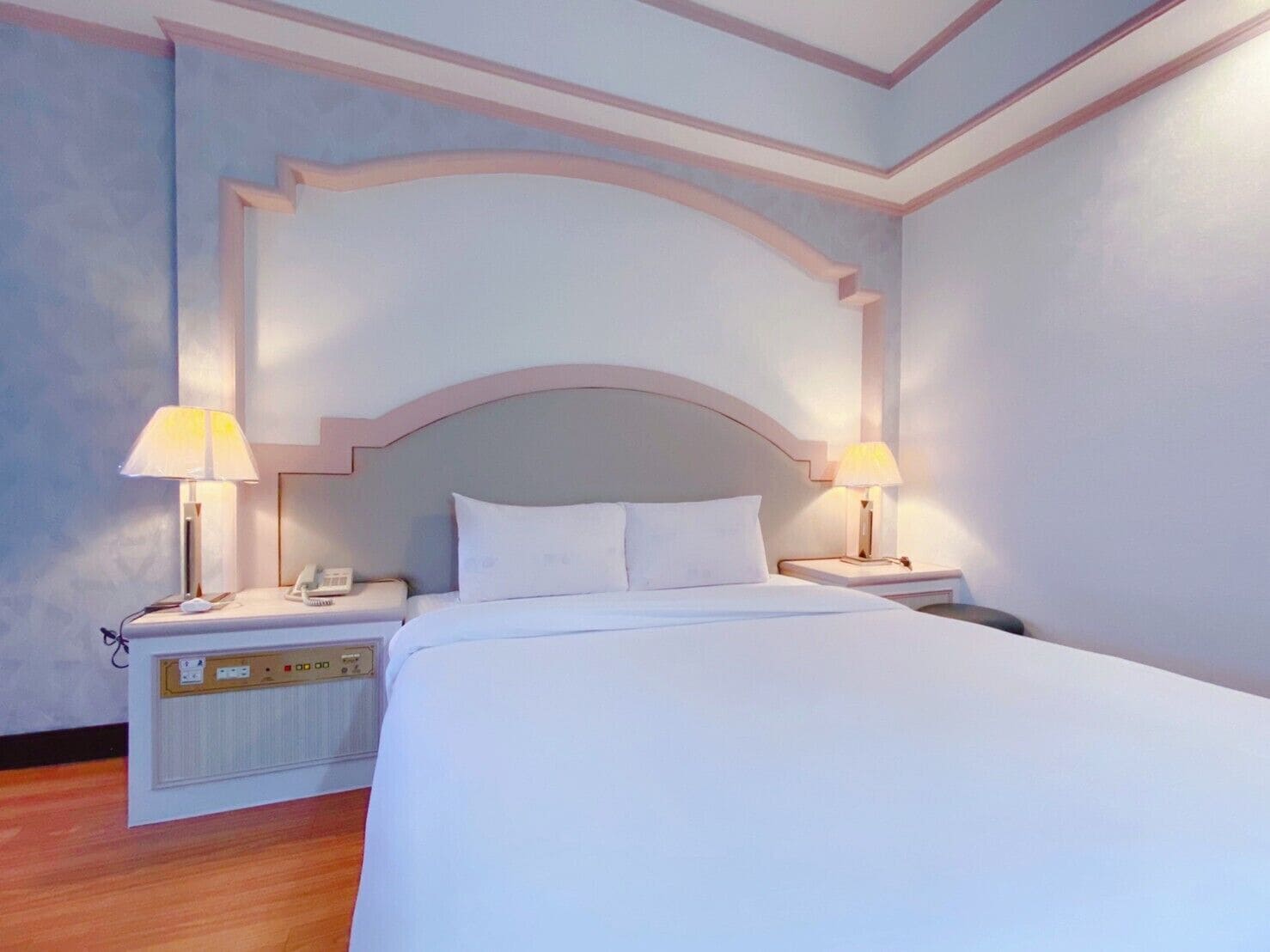Una habitación de hotel con una cama grande, dos mesitas de noche con lámparas, un atril con teléfono y un panel de control en la pared.