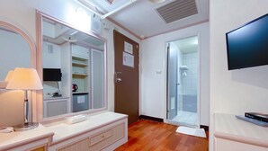 A hotel room with a wooden floor, a white vanity with a mirror, a flat-screen TV, and a door leading to a shower area.