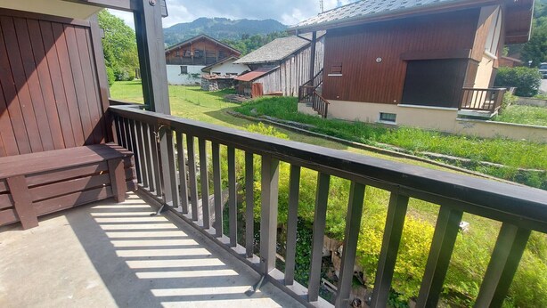Un balcon avec un banc en bois, une rambarde en métal et une vue sur une zone rurale avec des maisons et un ciel dégagé.