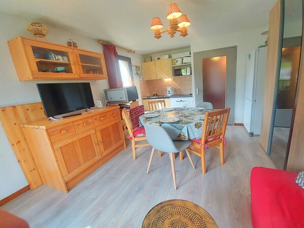 A compact kitchen with wooden cabinets, a dining table set for four, and a television on a wooden stand.