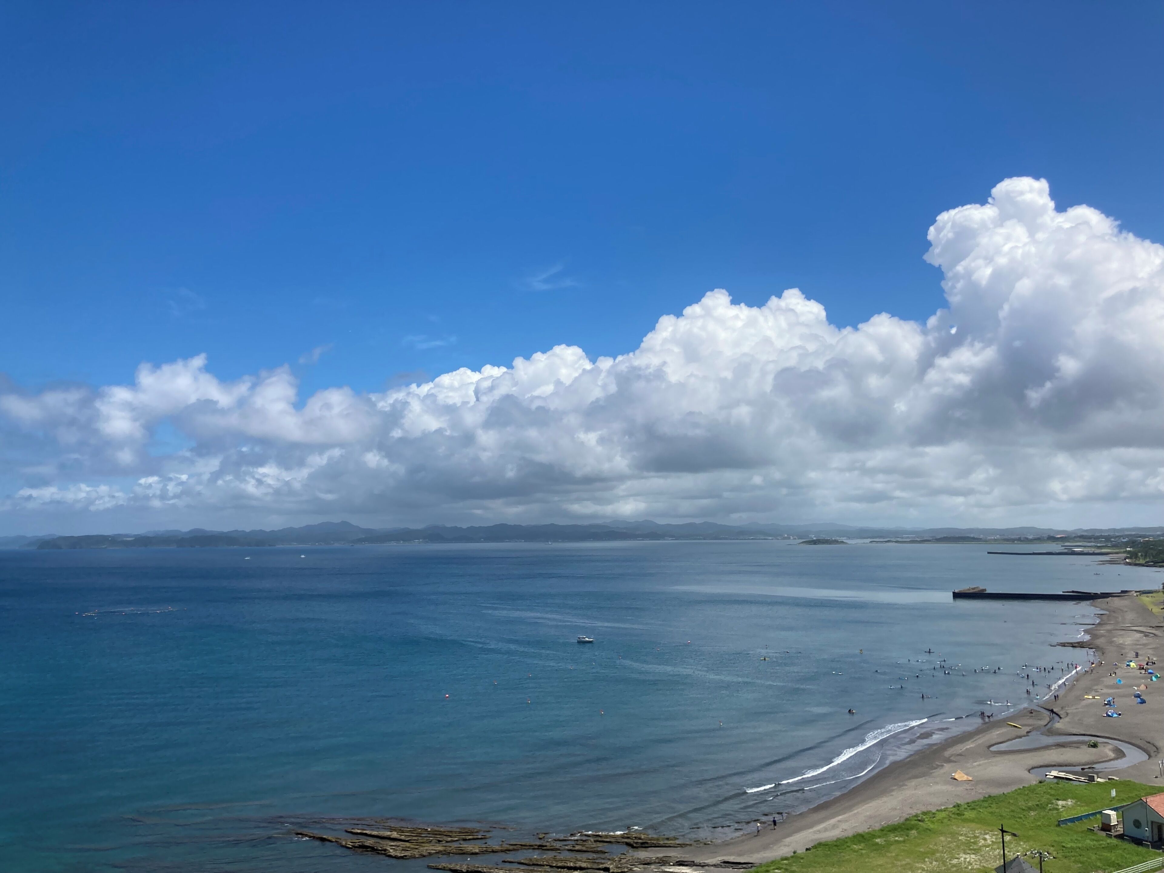 A coastal view with a beach, rocky shoreline, and distant mountains under a partly cloudy sky.