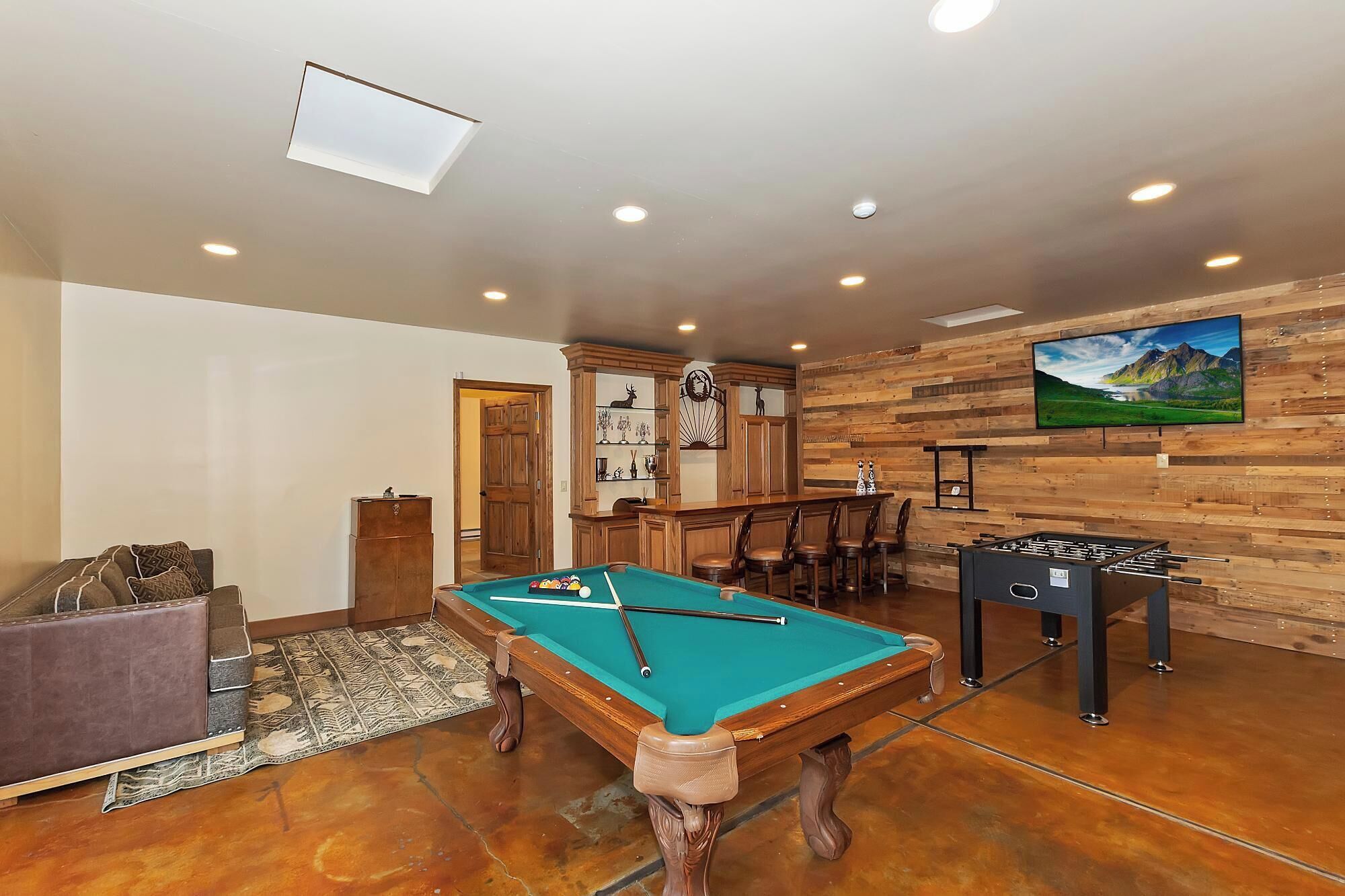 A pool table in a room with wooden paneling, a foosball table, and a bar area.