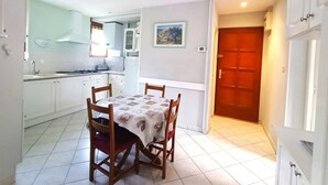 Une cuisine de taille réduite avec des meubles blancs, une table à manger avec des chaises et une porte rouge donnant sur une autre pièce.