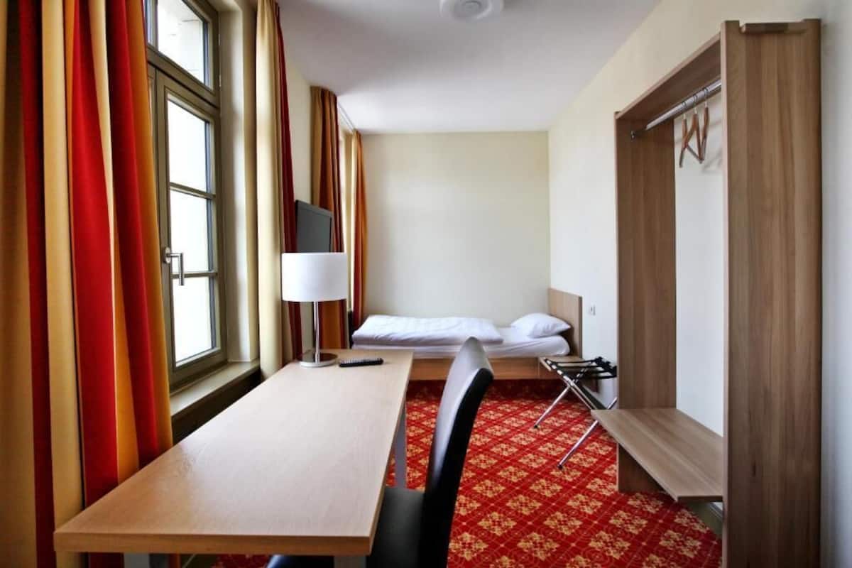 A hotel room with a wooden desk, a bed with white linens, a red patterned carpet, and a wooden wardrobe.
