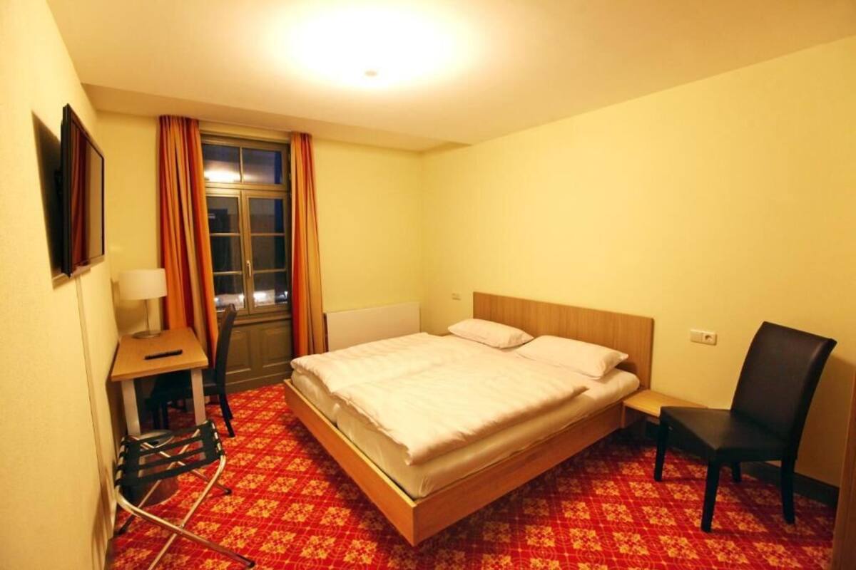 A hotel room with a single bed, a desk with a chair, a small table, and a window with curtains.