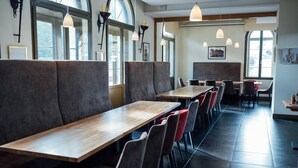 Ein Restaurant mit langen Holztischen, gepolsterten Sitzgruppen, großen Fenstern und Pendelleuchten.