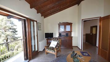 Un soggiorno con soffitto in legno, un'ampia finestra, televisore, una poltrona con motivo floreale, un mobile contenitore e un piccolo tavolino rotondo.