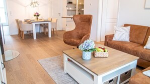 Ein modernes Wohnzimmer mit einem braunen Sessel, einem Couchtisch mit Blumen und einer braunen Couch. Im Hintergrund ist eine Essecke zu sehen.