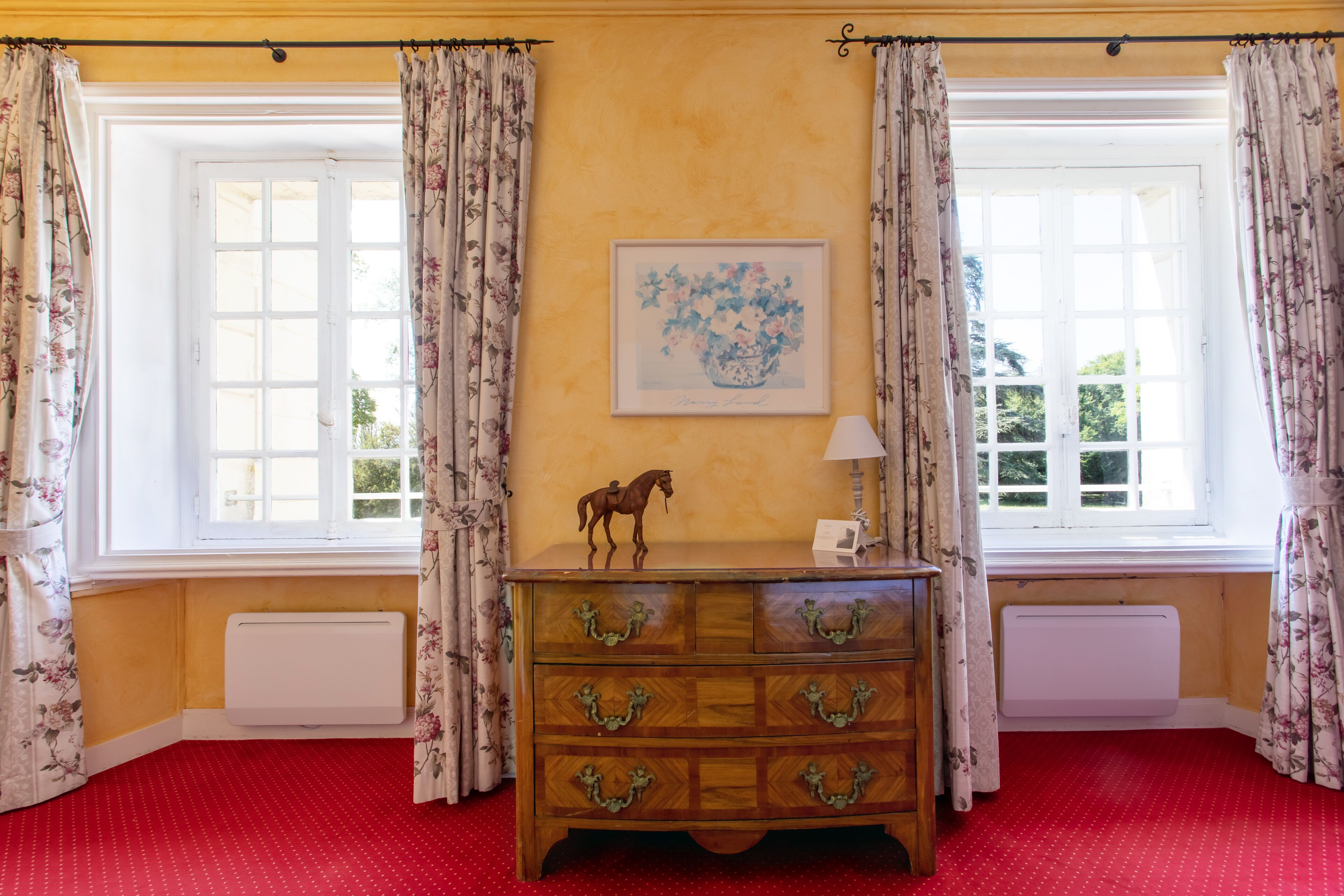 Una habitació amb un taulell de fusta, una obra d'art floral emmarcada i un radiador sota una finestra amb cortines florals.