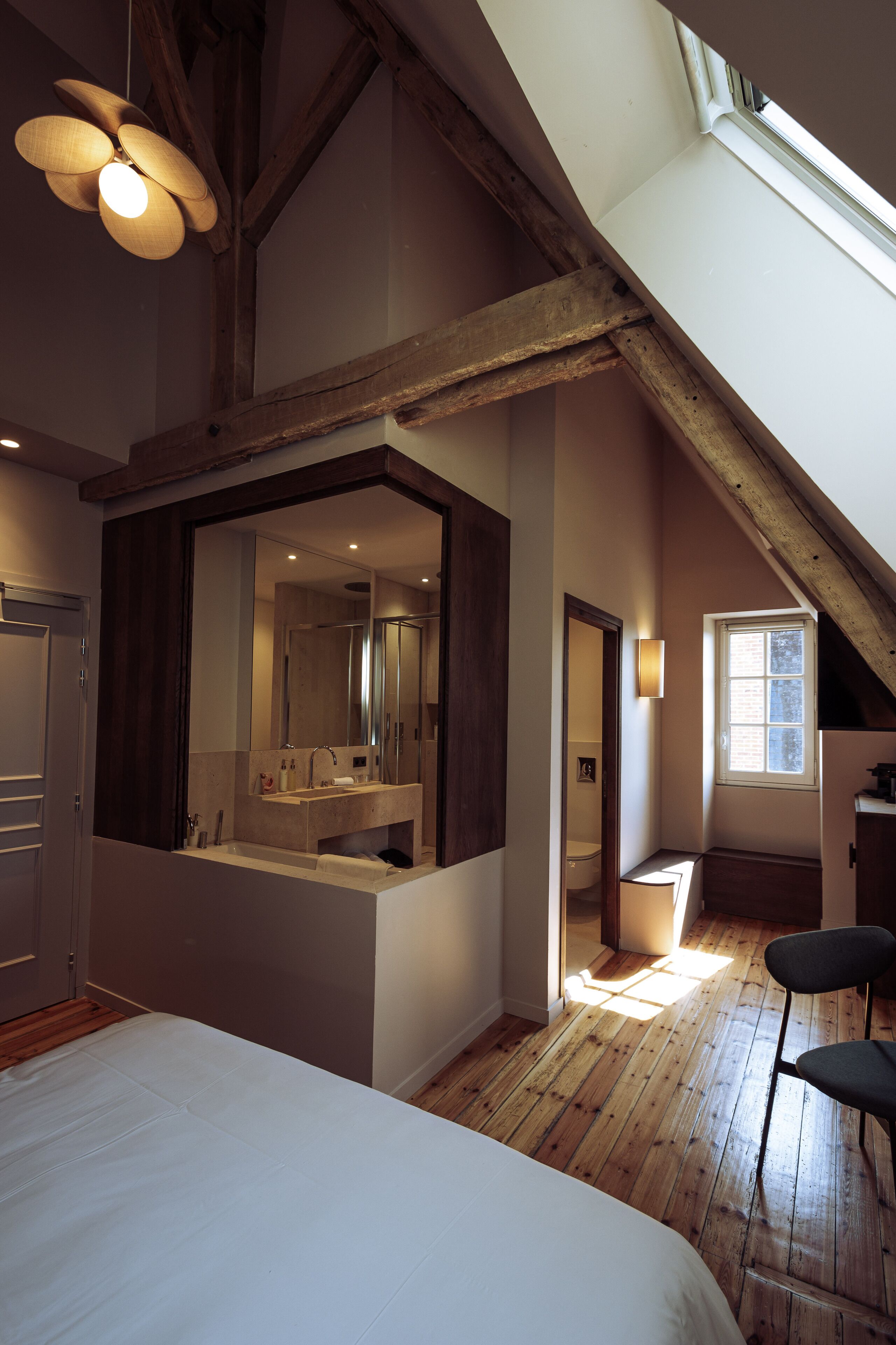 Een slaapkamer met een groot bed, houten plafond, dakraam, badkamer met spiegel en een raam.