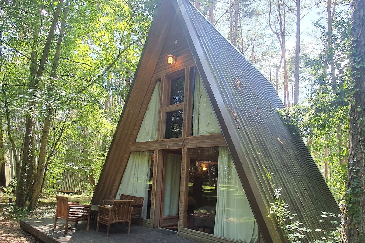 A wooden A-frame cabin with a deck and two chairs, surrounded by trees.