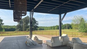 Un espace extérieur aménagé avec un coin salon, une pergola, une table avec des bouteilles et une vue sur un champ clôturé.