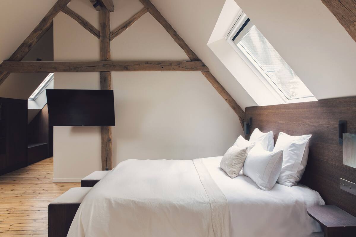 A bedroom with a sloped ceiling, wooden beams, a bed with white bedding, a bedside table, and a flat-screen TV mounted on the wall.