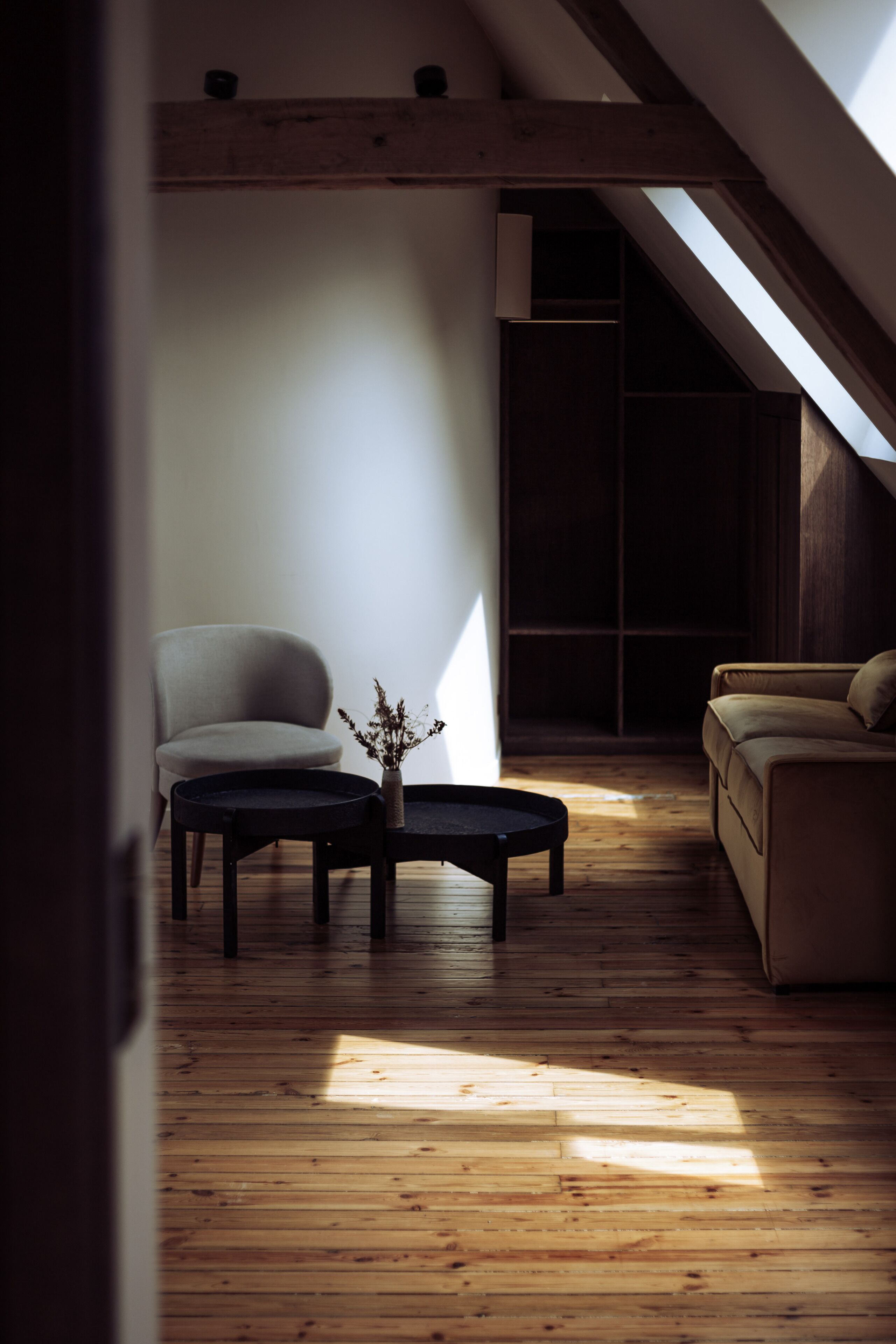 Una habitación con techo inclinado, suelo de madera, un sofá, una silla y una pequeña mesa con un florero.