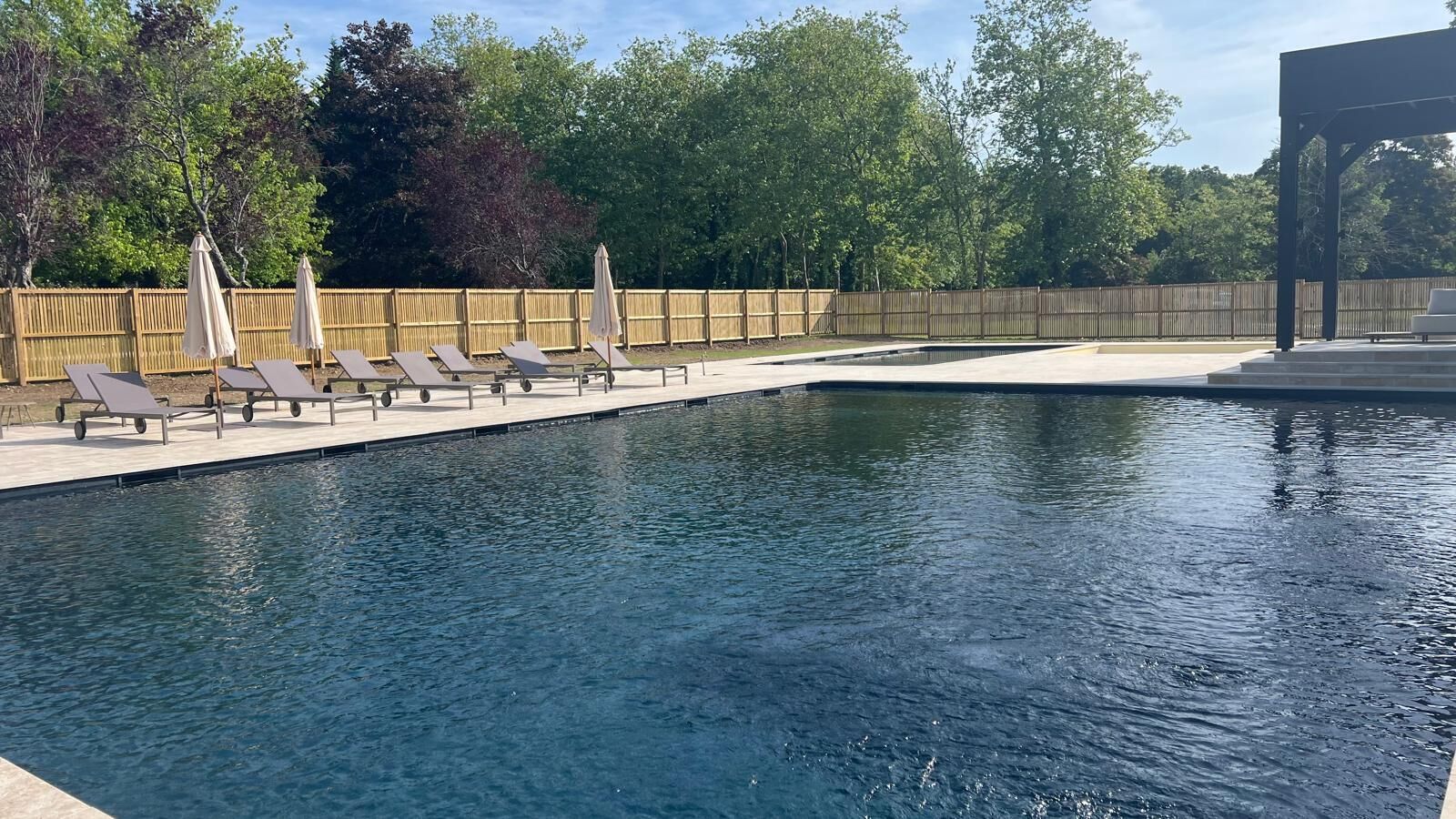 A swimming pool with lounge chairs and umbrellas, surrounded by a wooden fence and trees.